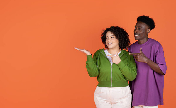 Special offer for two. Cute cheerful loving multiracial couple in colorful casual outfits embracing and pointing at free space on orange studio background, showing nice advertisement
