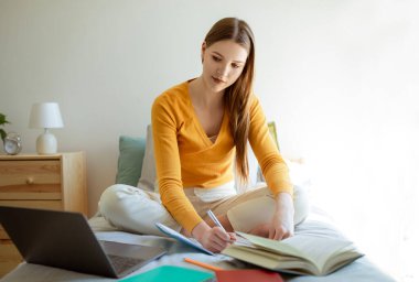Çevrimiçi Öğrenme. Genç kız öğrenci dizüstü bilgisayara bakıyor ve not alıyor, ödevlerini modern yatak odasında yapıyor. Uzaklık Çalışması ve E-Öğrenme Teklif Reklamı
