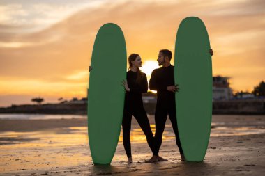 Günbatımında sahilde sörf tahtası olan mutlu milenyum çifti, dalış kıyafetleri giymiş romantik erkek ve kadın birbirlerine bakıyor ve eğleniyorlar, birlikte sörf yapıyorlar, uzay kopyaları yapıyorlar.