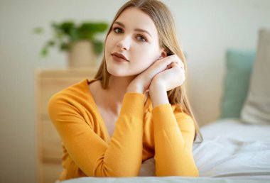 Uzun sarı saçlı sakin bir kadının portresi günlük kıyafetlerle poz veriyor, elleri yüze yakın, modern yatak odasında oturuyor. Güzel Öğrenci Kadın 'ın vesikalığı. Doğal Güzellik Konsepti