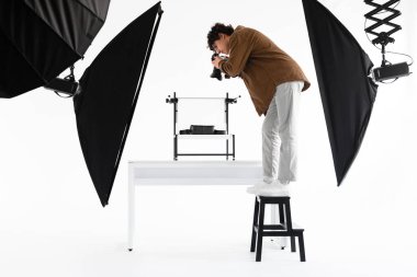 Genç adam, merdivenin üzerinde duruyor ve çantaların yüksek açılı fotoğraflarını çekiyor, profesyonel fotostudio 'da çeşitli ekipmanlarla çalışıyor, yan görüş