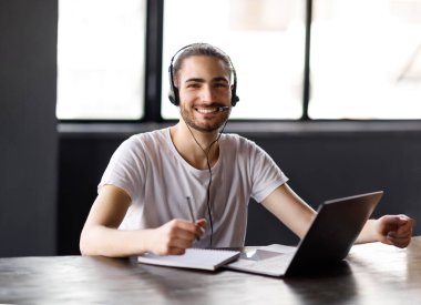 E-öğrenme. Modern ofis içinde masada otururken kulaklık takan, kameraya gülümseyen dizüstü bilgisayar pozu veren mutlu Orta Doğulu adam. Çevrimiçi Genç İşadamı Öğreniyor ve Çalışıyor