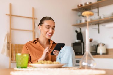 Yemek yerken, masada otururken ve telefonda mesajlaşırken, sosyal ağlara göz atarken ya da online alışveriş yaparken, mutfakta akıllı telefon kullanan heyecanlı bir bayan.