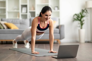 Çevrimiçi egzersiz programları. Güzel esmer, uzun saçlı, spor kıyafetli genç bayan yoga minderinde, egzersiz yapıyor, dizüstü bilgisayar kullanıyor, fitness videosu izliyor, mekanı kopyalıyor.