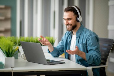 Video çağrısı. Kulaklıklı neşeli genç iş adamı ofiste telekonferans yapıyor, gülümseyen milenyum erkek girişimcisi iş ortağıyla çevrimiçi görüşme için dizüstü bilgisayarını kullanıyor,