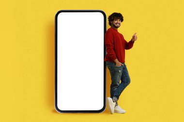 Güzel bir mobil uygulama. Olumlu, neşeli, genç, Hintli bir adam. Beyaz ekranlı, akıllı bir telefonun başında, sarı arka planda izole edilmiş bir şekilde duruyor.