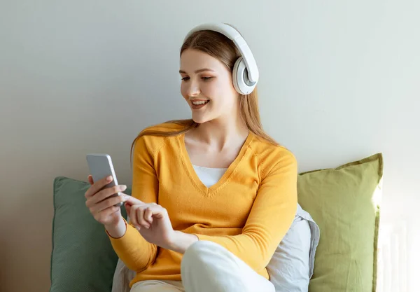デジタル ファン 現代の寝室の屋内でソーシャルメディアで音楽やテキストをオンラインで聞くスマートフォンを使用してイヤフォンを持つ若い女性 ベッドの上に座って ガジェットレジャーコンセプト — ストック写真