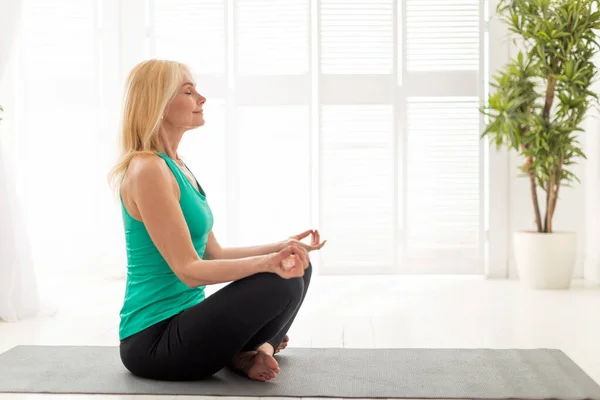 Wellness Concept. Smiling Senior Lady Meditating On Yoga Mat At Home, Portrait Of Calm Relaxed Mature Woman Sitting In Lotus Position, Practicing Meditation In Light Room, Side View With Copy Space