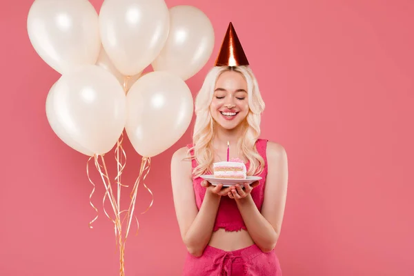 帽子のきれいな女性はケーキを持っていて ピンクの背景 スタジオに隔離された膨脹可能な風船の近くにポーズをとって 誕生日の願いを作ります 祝日からの感情 — ストック写真