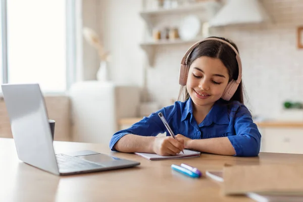 在线教育 在耳机里快乐的阿拉伯小女孩 带着笔记本电脑学习 记笔记 在家里的办公桌边写作业 男校女生远程学习 — 图库照片