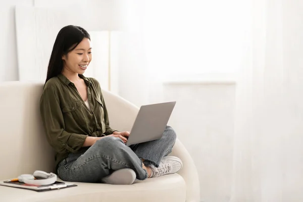 彼の膝のコンピュータノートパソコンとソファの上に座ってカジュアルな服を着て美しい千年紀の中国の女性の笑顔 Pcのキーボードで入力 ホームインテリア コピースペース チャットボット 日常生活のAi — ストック写真