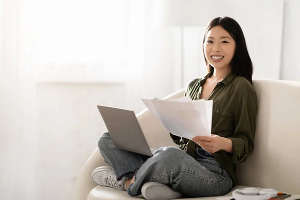 幸せな陽気な若いアジアの女性のビジネスコンサルタントは 独立した請負業者の自宅から 彼女の膝の上にコンピュータのラップトップでソファに座って 文書を保持し スペースをコピーします リモートジョブ — ストック写真