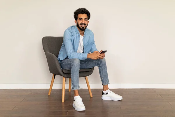 Vrolijke Jonge Indiase Man Met Smartphone Handen Ontspannen Een Fauteuil — Stockfoto