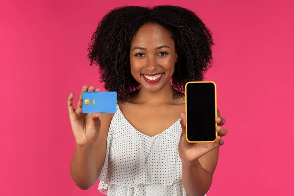 明るく若い黒の巻き女性は ピンクの背景 スタジオに隔離されたオンラインショッピングのための空の画面でクレジットカードと電話を示しています 金融アプリ キャッシュバック ウェブサイト 広告やオファー — ストック写真