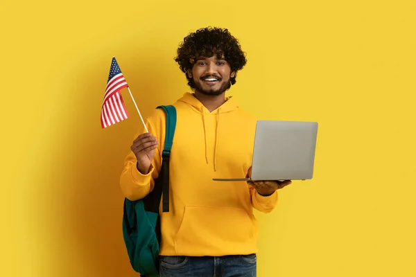 幸せな若い東アジアの男 バックパックを運ぶ黄色のパーカーを着て 米国のノートパソコンやフラグを保持する学生の後ろに黄色の背景に孤立した 海外での教育 アメリカの概念で学ぶ — ストック写真