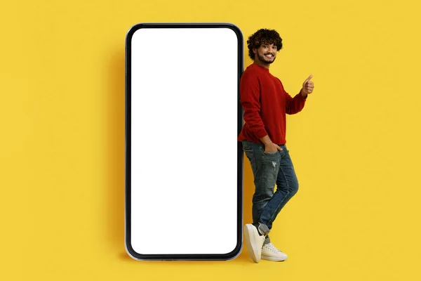 有趣的移动应用程序 穿着休闲装 快乐而又英俊的印度小伙子 站在白色空白屏幕的大智能手机旁 露出大拇指 在黄色的背景上被隔离 模仿着 — 图库照片