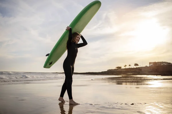 stock image Portrait of young female surfer walking with surfboard at beach on sunset, happy millennial woman wearing wetsuit going out of sea after surfing, enjoying water sports and active lifestyle