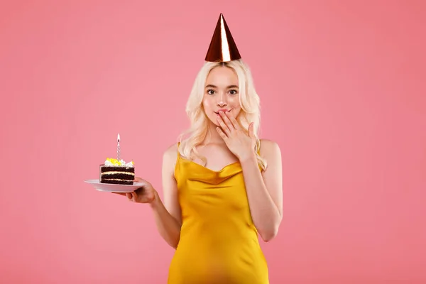 ハッピー喜び女の子でパーティー帽子保持ピースのケーキと閉じ口とともに手 誕生日を祝います 上のピンクのスタジオの背景 — ストック写真