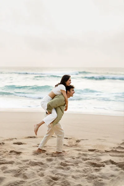 全长拍摄快乐的年轻人在海滩上搭便车给他的女朋友 夫妻在海岸线上享受约会和时间 垂直拍摄 — 图库照片