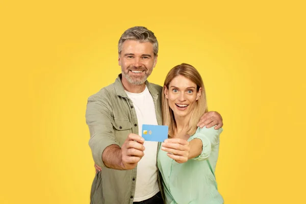 Easy digital payment. Excited european middle aged spouses hugging, showing credit card and smiling at camera, recommending banking service over yellow studio background