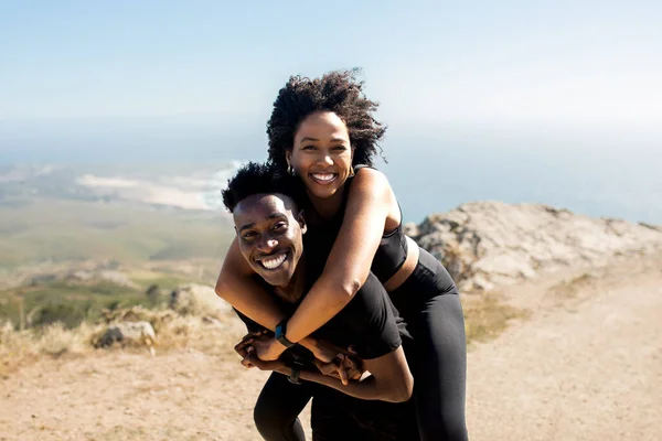 Excited african american couple resting during workout outdoors on rocks at ocean, man giving piggyback ride to lady. Love, health care, active lifestyle and sports outside