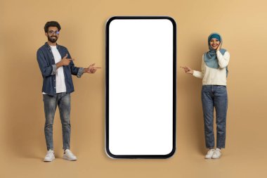 Harika bir tanıtım. Heyecanlı İslami Çift Büyük Boş Telefon Ekranını Gösteriyor, Yeni Mobil Uygulama veya Web Sitesindeki Mutlu Arap Adam ve Kadın, Bej Arkaplanda Poz Veriyor, Mobilya, Kolaj