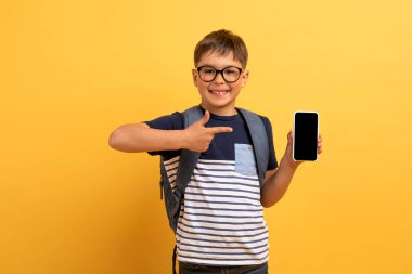 Eğitici mobil uygulama. Mutlu okul çocuğu beyaz giysili, gözlüklü ve sırt çantalı siyah beyaz ekranlı, sarı arka planda izole edilmiş modern akıllı telefonu gösteren sevimli bir çocuk.