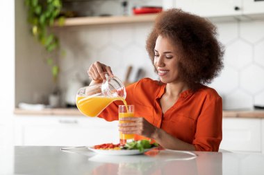 Güzel siyahi kadın portresi evde sağlıklı kahvaltı yapıyor, mutlu Afro-Amerikan bayan masada oturuyor, lezzetli yemeklerin tadını çıkarıyor ve portakal suyunu sürahide bardağa dolduruyor,