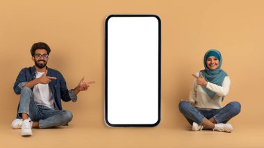 Mutlu Müslüman çift oturuyor ve boş ekranlı büyük akıllı telefonu işaret ediyor, yeni uygulama öneriyor, tesettürlü neşeli Arap erkek ve kadın bej arka planda cep telefonuyla poz veriyor