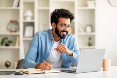 Çevrimiçi Eğitim. Genç Hintli adam dizüstü bilgisayarda giyiniyor ve not alıyor, gülen doğu milenyumu çocuğu uzaktan öğrenmenin keyfini çıkarıyor, İçişleri Bakanlığı 'nda masa başında oturuyor, boş yer var.