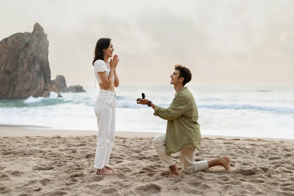 stock image Romantic proposal on the seashore. Loving young man with engagement ring making proposal to happy woman on beach on coastline, side view