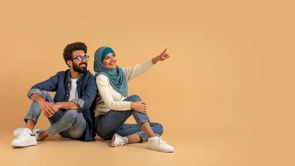 いい提案だ ムスリムカップルが床に座り コピースペースで脇を指差して笑顔 ビージスタジオの背景に広告のための無料の場所を示すヒジャブで幸せなアラビア人男性と女性 パノラマ — ストック写真