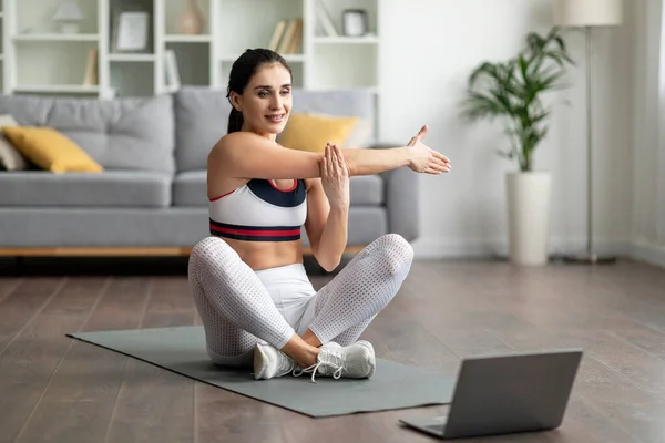穿着白色运动服的健康而又漂亮的年轻女子坐在健身垫上 伸展双臂 在家里练习瑜伽 在网上观看视频 跟随教练 复制空间 — 图库照片