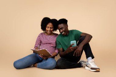 Telefonu olan genç Afro-Amerikalı bir koca, koca göbekli hamile bir eş not defterine not tutuyor, bej renkli stüdyo arka planında izole edilmiş. Bebek için birlikte uygulama, sevgi, destek, aile