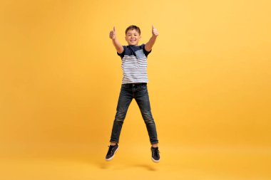 Sevimli neşeli, neşeli okul çağındaki çocuk havada zıplayan ve kameraya başparmak kaldıran, sarı stüdyo arka planında izole edilmiş, tam boy, kopyalama alanı öneren.