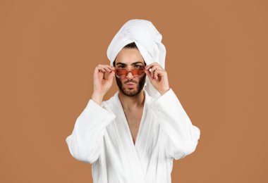 Erkek özgüveni. Beyaz bornoz giyen, kalp şeklinde güneş gözlüğü takan, cilveli dudakları olan, kahverengi arka planda duran güzelliğini önemseyen Arap bir adam. Stüdyo Portresi