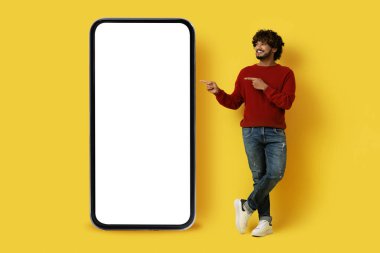 Chatbot, çevrimiçi asistan. Şık giyinmiş, şık giyinmiş, sarı arka planda izole edilmiş beyaz ekranlı büyük bir cep telefonunu işaret eden mutlu, yakışıklı, doğulu adam.