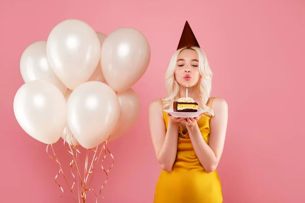 誕生日の帽子と風船がケーキの上にキャンドルを吹いている可愛い女性 ピンクのスタジオの背景に休日のパーティーやお祝いを持っている フリースペース — ストック写真