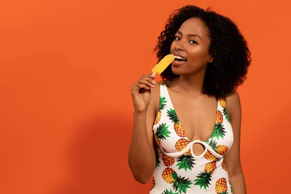 stock image Summer mood concept. Cheerful relaxed hot sexy attractive young black woman wearing trendy colorful swimsuit having delicious ice cream, chilling on orange studio background, copy space
