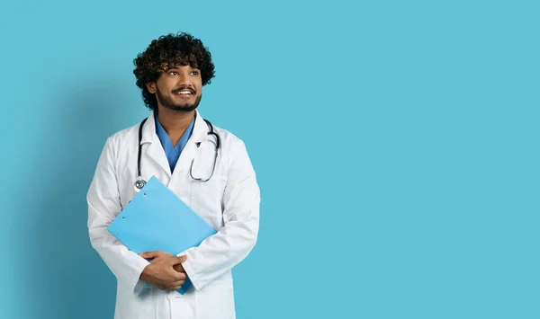 他手里拿着病历 笑容满面 卷曲胡子的印度年轻医生 看着诊所或医院广告的复制空间 背景是蓝色的 — 图库照片