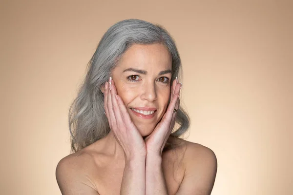 一个性感的 年长的 灰头发的女人的画像 在镜头前触摸着她的脸 微笑着 摆出一副米色工作室的样子 享受温泉治疗的肌肤柔软的老太太 — 图库照片