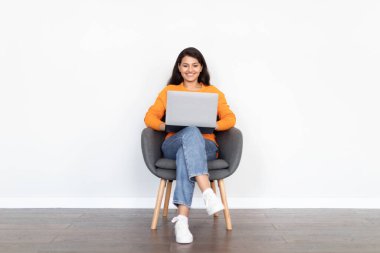 Online asistan, sohbet robotu, günlük hayatta yapay zeka. Gülümseyen genç Hintli kadın günlük kıyafetler giyiyor bilgisayar bilgisayarını beyaz duvar arkasında kullanıyor, boşluğu kopyalıyor.