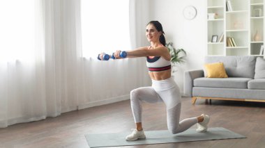 Zinde kalmak, spor yapmak, kadınlar için evde egzersiz yapmak. Formda, motive olmuş, gülümseyen, beyaz spor kıyafetli, halterleri kaldıran, fotokopi alanı, iç mekan, panorama...