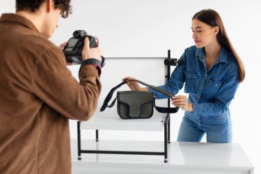 Erkek fotoğrafçı ve smm yöneticisi modern fotostudio 'da beyaz platform arka planında şık bir el çantasının fotoğraflarını çekiyor. Kadın içerik fotoshoot yapımına yardım ediyor.