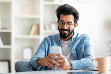 Genç Hintli erkek Freelancer İç İşleri 'nde çalışırken akıllı telefon kullanıyor, Mutlu Doğulu adam cep telefonuyla mesaj atıyor ya da yeni uygulamalara bakıyor, modern teknolojilerin tadını çıkarıyor, Space' i kopyalıyor.