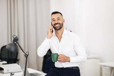 Beyaz tişörtlü, sakallı, mutlu Avrupalı bir yetişkin telefonda konuşuyor. Ofiste birlikte çalışırken bir fincan kahve içiyor. Müşteriyle iş, iş ve profesyonel iletişim