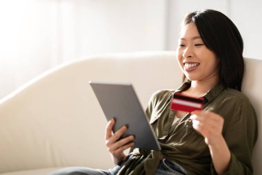 Sanal bankacılık, e-ticaret. Kanepeye yaslanmış, dijital tablet ve banka kartı tutan, online bankacılık, fotokopi çekilen sıradan giysili neşeli, genç Asyalı kadının yakın çekimi.