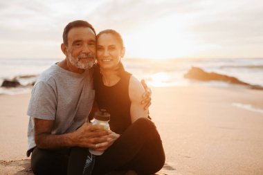 Sağlık, spor ve aşk. Mutlu Kafkasyalı kadın ve erkek spor kıyafetleri içinde bir şişe suyla kucaklaşır, eğitimin tadını çıkartır, plajda, dışarıda, dışarıda, güneş parlamasıyla aktif bir yaşam tarzı sürdürür.