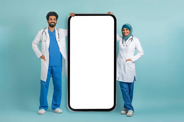 阿拉伯女医生和男医生身着制服 站在白色屏幕的空白大智能手机旁边 穆斯林医生们展示着医疗广告的复制空间 摆出一副蓝色背景 模仿着 — 图库照片