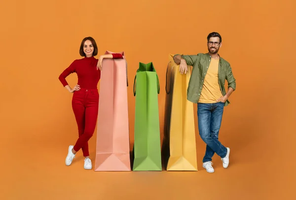 销售旺季 快乐的购房者们靠在大纸袋上 在摄影棚里站在橙色的背景下 享受广告折扣和微笑 大合院 全长一枪 — 图库照片
